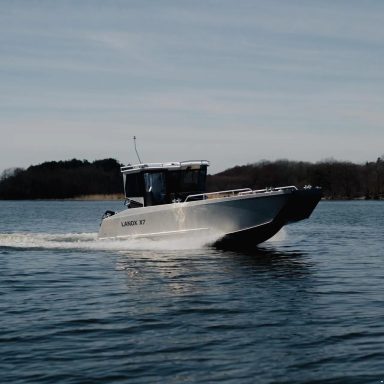 Ein Schnellboot fährt über ruhiges Wasser, mit Bäumen im Hintergrund.