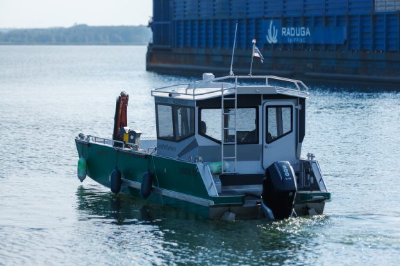 Ein grünes Boot fährt ruhig im Wasser, vor einem blauen containerbeladenen Hafen.