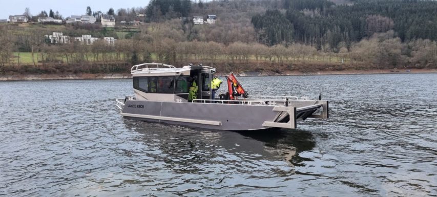 Ein Boot mit zwei Personen fährt auf einem ruhigen Gewässer zwischen bewaldeten Hügeln.