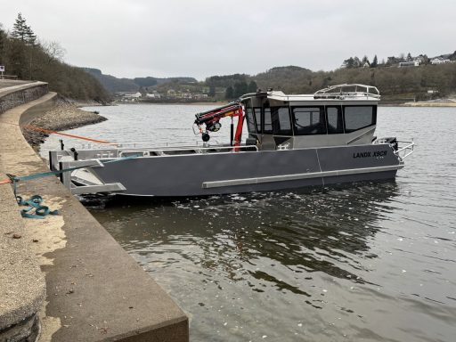 Ein grauer Bootstyp am Ufer eines ruhigen Gewässers mit einer Landschaft im Hintergrund.
